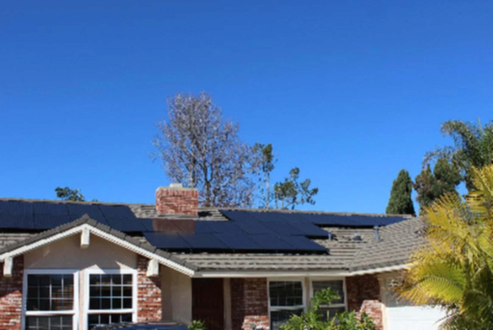 Single-story California home with rooftop solar panels, a brick chimney and palm trees under a clear blue sky